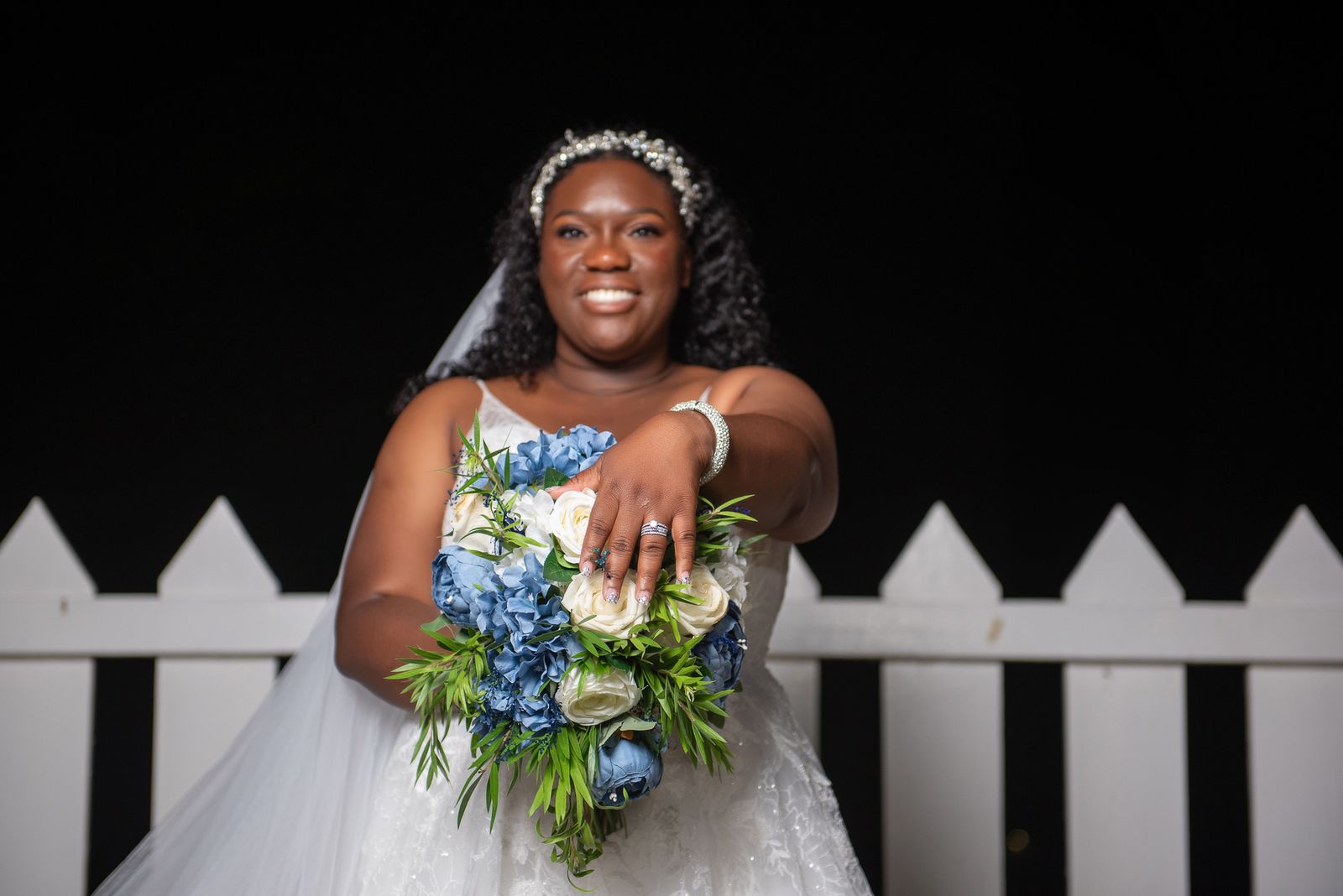 The bride holding a blue and white bouquet against a white picket fence at night