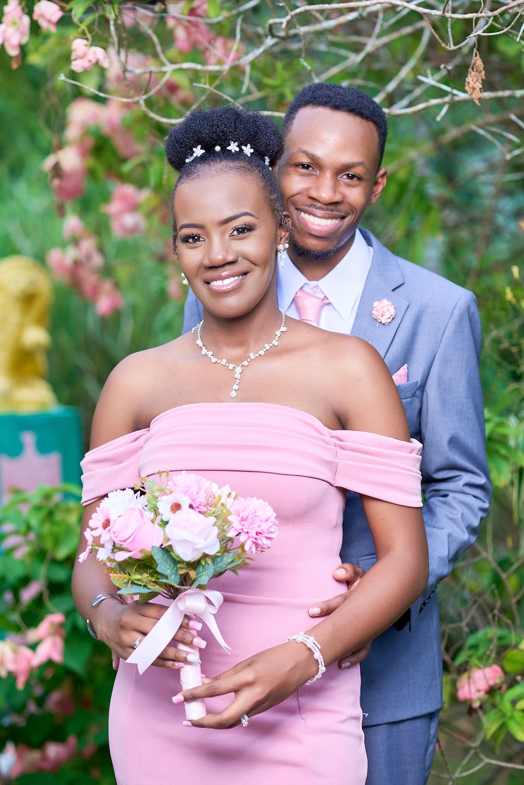 A couple in pink and grey among flowering shrubs, her bouquet held between them