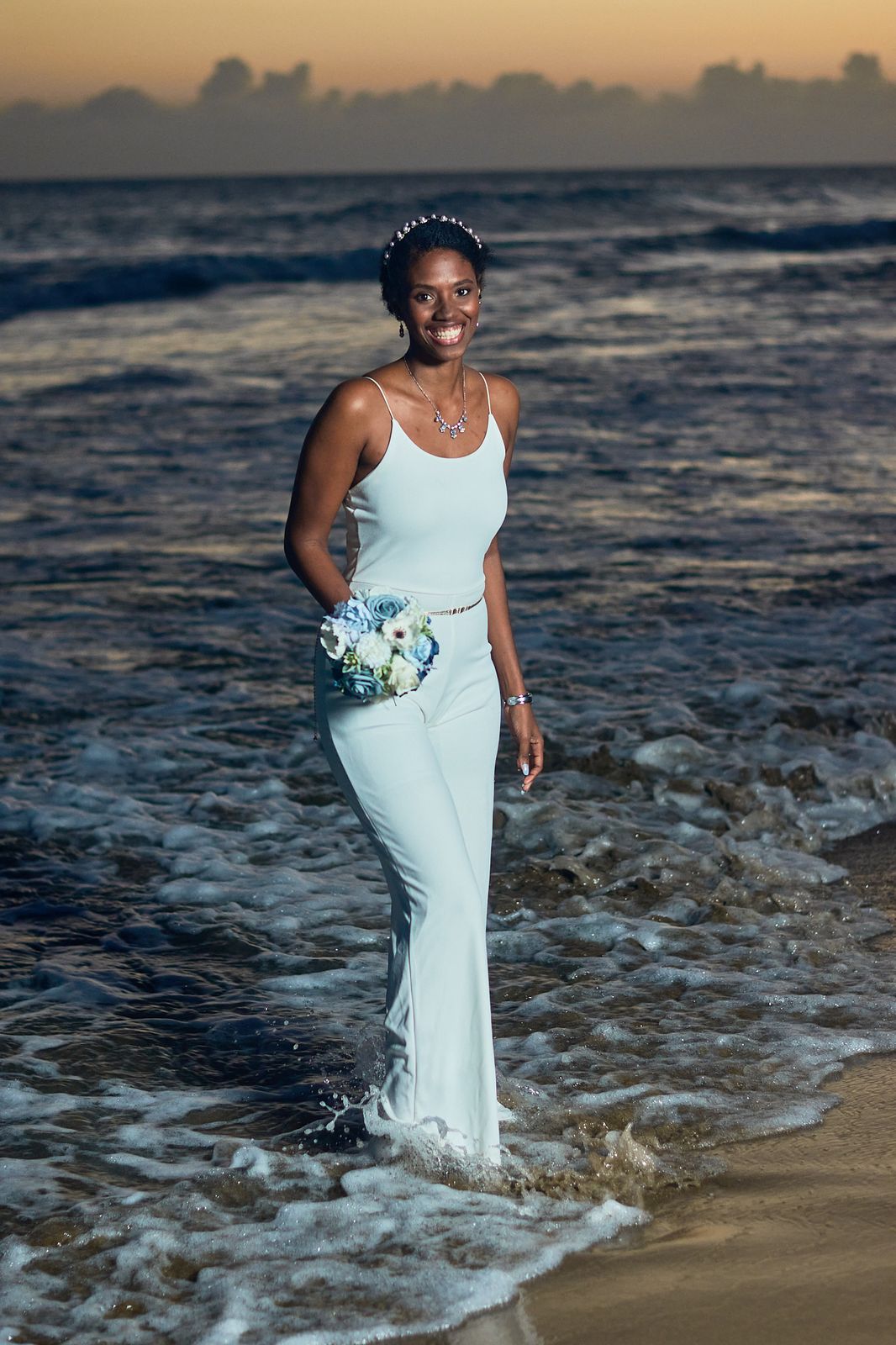 The bride standing in the shallow surf at dusk, holding a pale bouquet