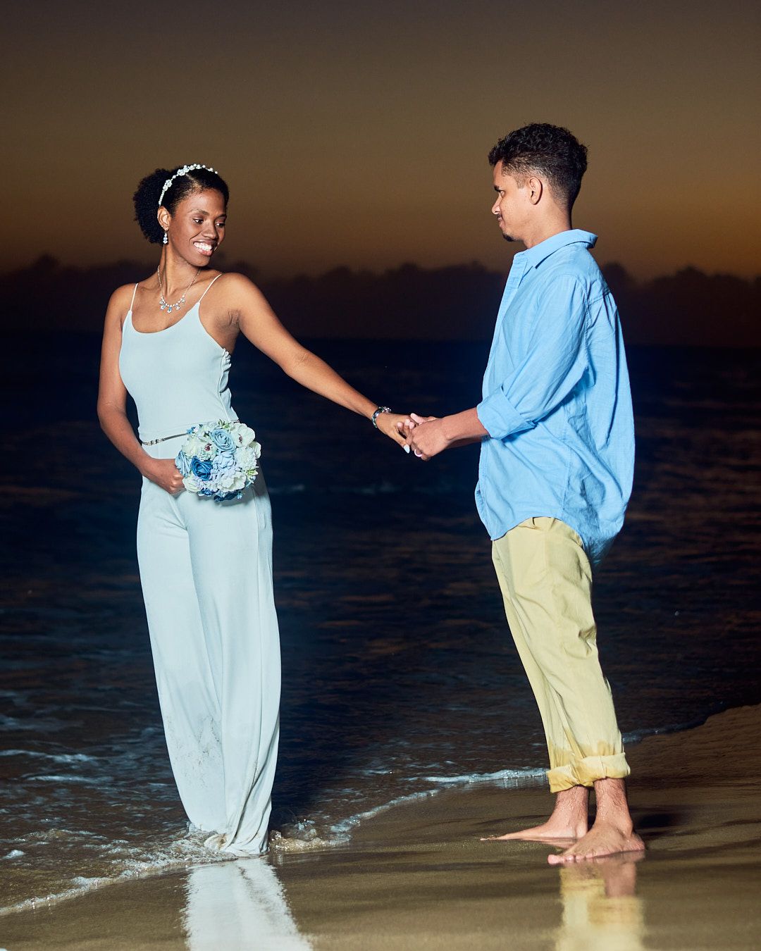 The couple holding hands at the water’s edge as the sun goes down